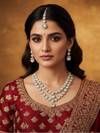 Woman wearing traditional jewelry and a red saree with gold patterns against a warm brown background